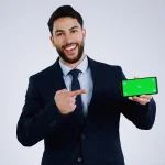 Professional businessman in dark suit pointing at smartphone with green screen display, presenting MyGreenBucks financial app against gray studio background