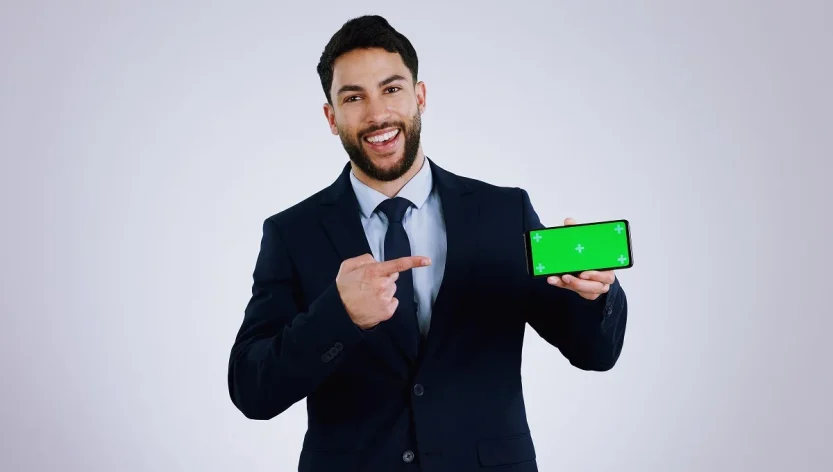 Professional businessman in dark suit pointing at smartphone with green screen display, presenting MyGreenBucks financial app against gray studio background